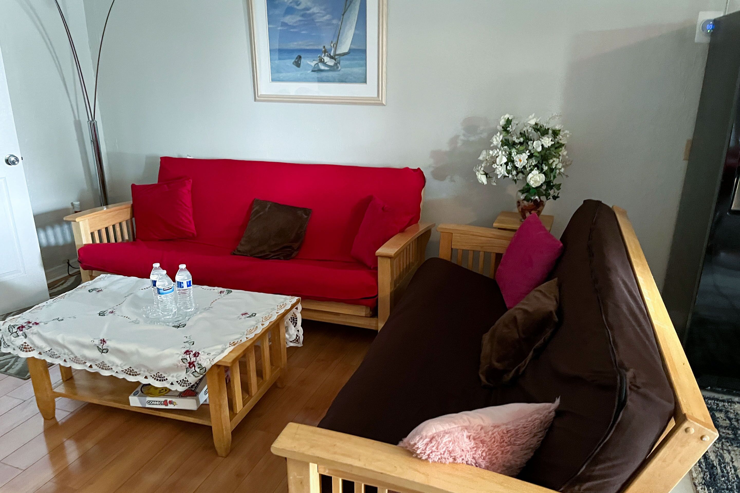 Una sala de estar con un sofá rojo, un sofá marrón, una mesa de centro de madera con mantel floreado y un jarrón con flores blancas.