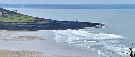A coastal landscape with a sandy beach, rocky shoreline, and a grassy hill with a building.
