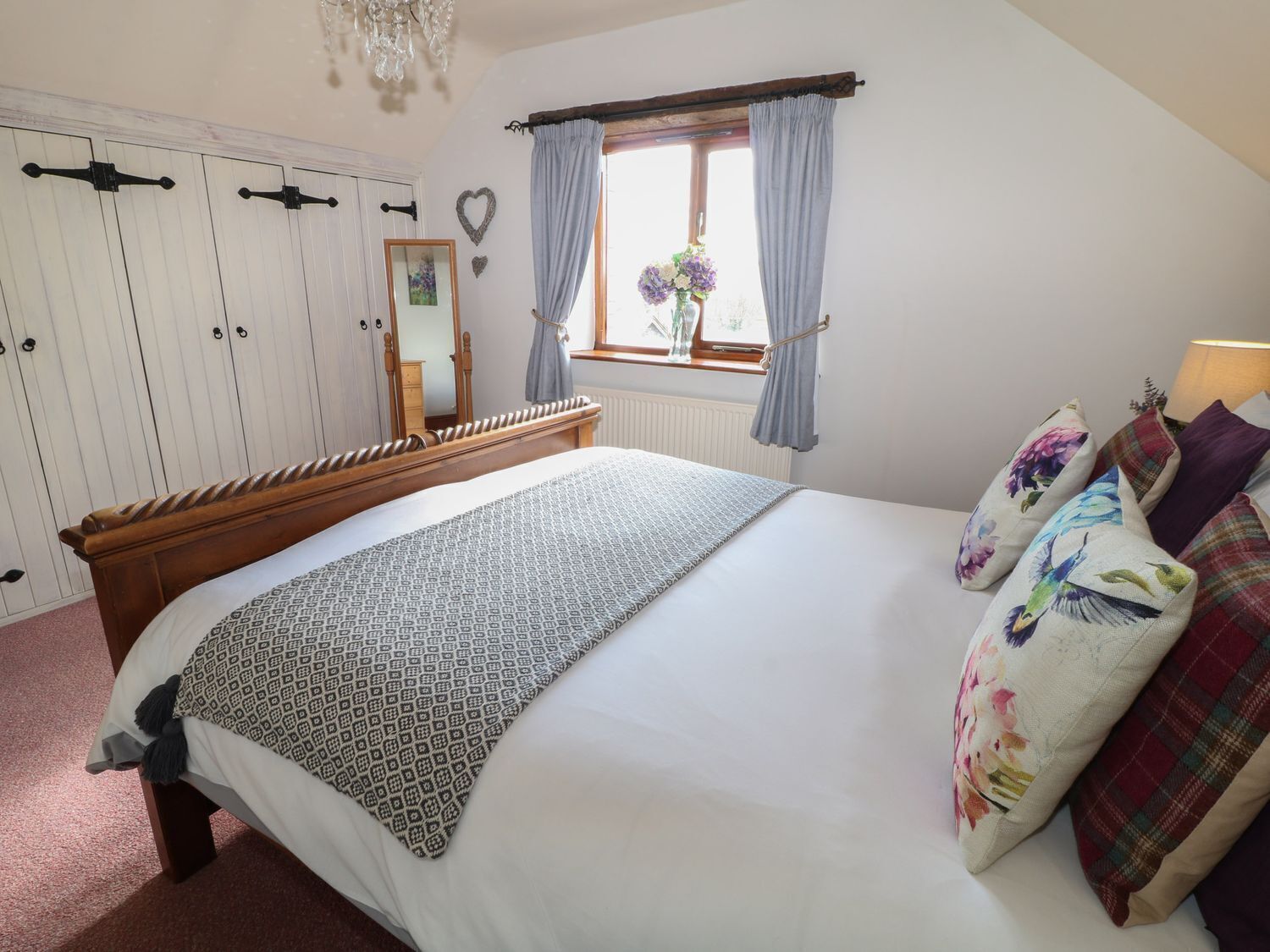 Une chambre avec un lit en bois, une fenêtre avec des rideaux, un radiateur et une armoire décorée d’un motif en forme de cœur.