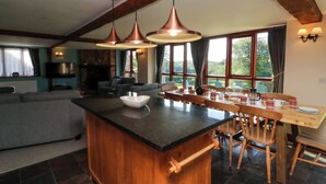 A spacious kitchen with a central island, wooden beams, large windows, and a dining area with wooden chairs and a table. - Apple Barn (South Molton)
