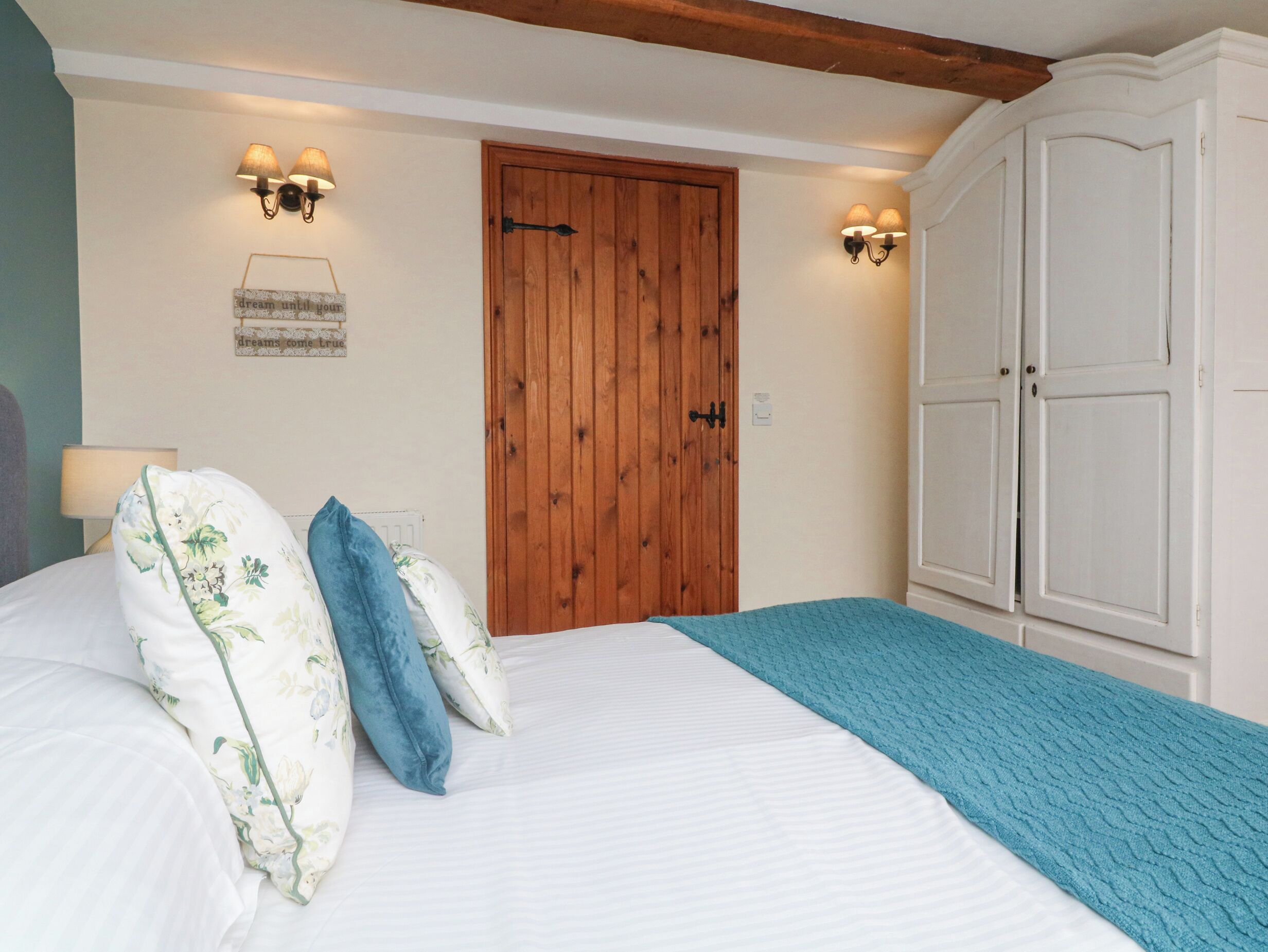 A bedroom with a wooden door, two wall-mounted lamps, a bed with floral pillows, and a blue blanket.