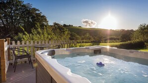 A hot tub with a wooden deck and a white fence, surrounded by greenery and a clear sky. - Apple Barn (South Molton)