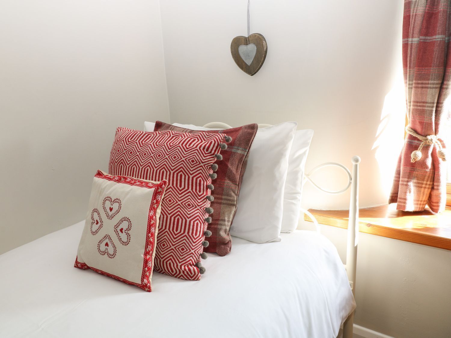 Un dormitorio con cama, almohadas, un adorno con forma de corazón en la pared y una ventana con cortinas.