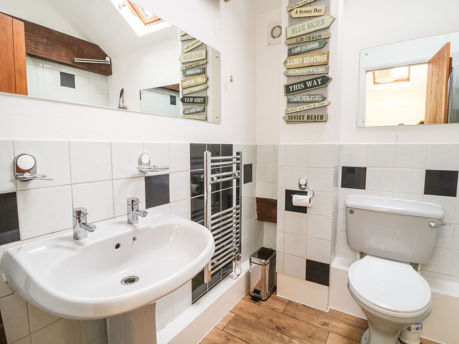 A modern bathroom with a white sink, a toilet, and a towel rack. There are black and white tiles on the walls and a skylight above the sink.
