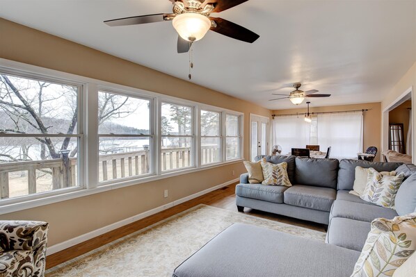 A spacious living room with large windows, a ceiling fan, and a gray sofa with patterned cushions.