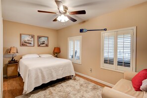 A bedroom with a bed, a ceiling fan, two windows with shutters, and a nightstand with a lamp.