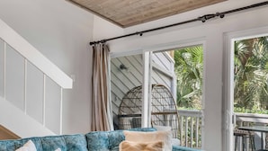 Quarto com sofá azul, mesa de centro de madeira, teto com vigas de madeira e uma janela grande com cortinas.