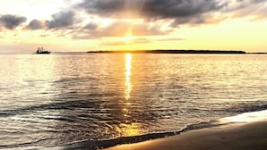 Um pôr do sol sobre um mar calmo, com um navio ao longe e uma praia em primeiro plano.