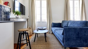 Una sala de estar con un sofá azul, una mesita redonda, un televisor montado en la pared y una planta en maceta.