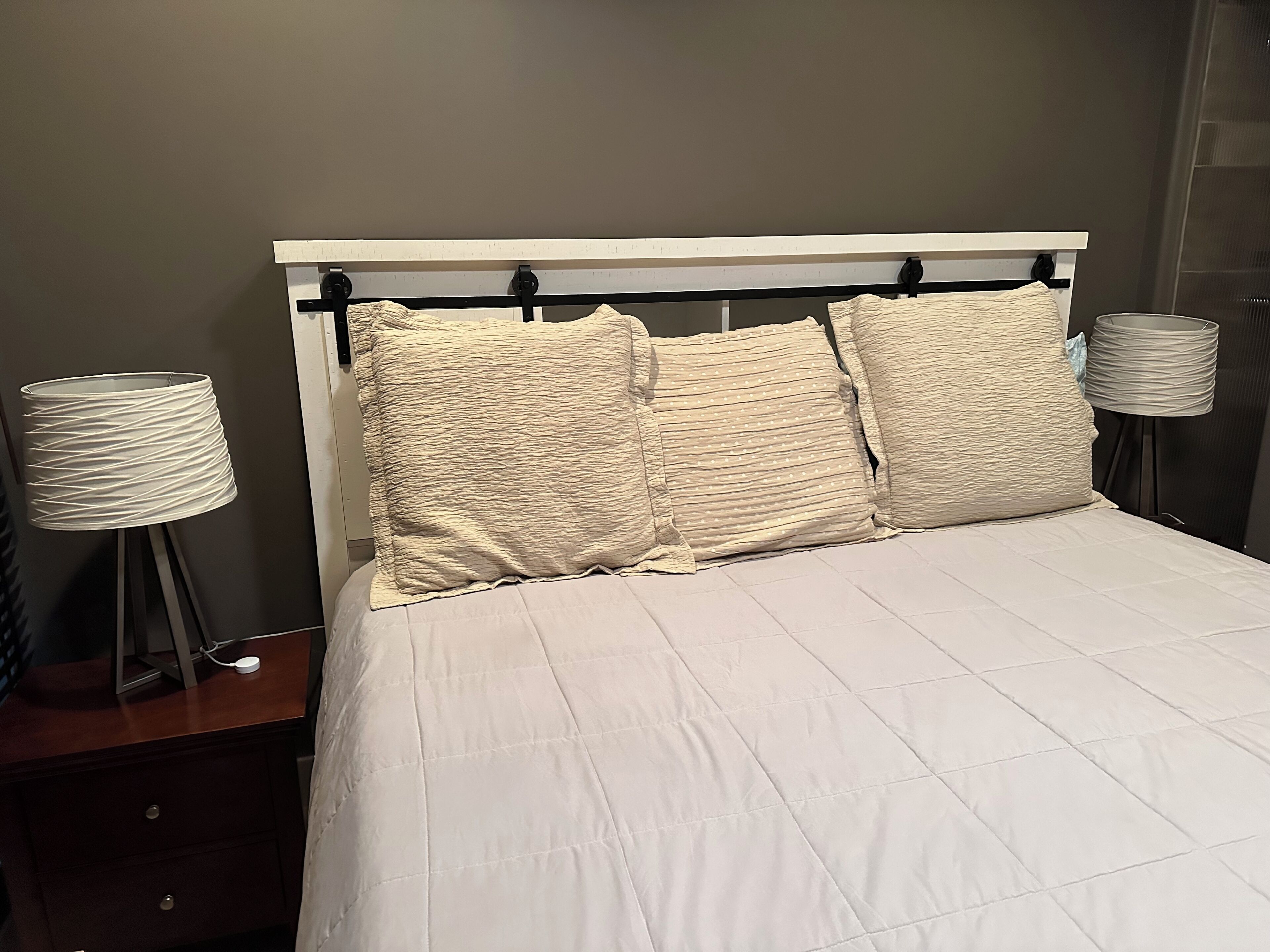 A bed with a white quilted bedspread and three textured pillows. There is a bedside table with a lamp on it.