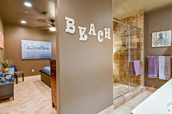 Ein Zimmer mit einem 'Beach'-Schild, einem Bett, einem Schreibtisch mit Pflanze und einem Duschbereich mit Glastür.
