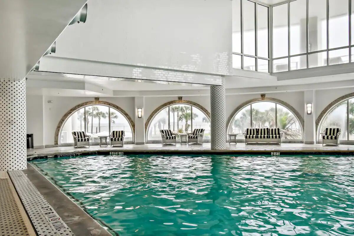 Une piscine intérieure à l’eau claire, entourée de fenêtres en arc de cercle et de chaises à rayures.