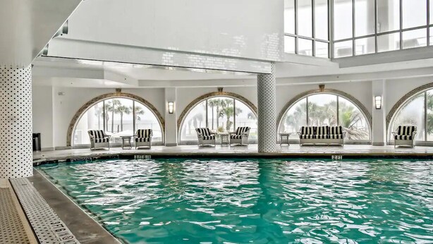 Una piscina interior con agua cristalina, rodeada de ventanas en arco y sillas a rayas.