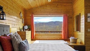 A wooden cabin bedroom with a bed, pillows, a nightstand, a window with curtains, and a view of a forested landscape. - Great View, Location, Studio Log Cabin, Hot Tub! (Gatlinburg)