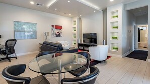Una sala de estar moderna con una mesa de comedor de vidrio, sofás blancos y negros, un televisor de pantalla plana y una pequeña zona de cocina con microondas y cafetera.