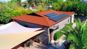 Una casa con techo rojo, paneles solares, un patio con sombra y un jardín con palmeras.