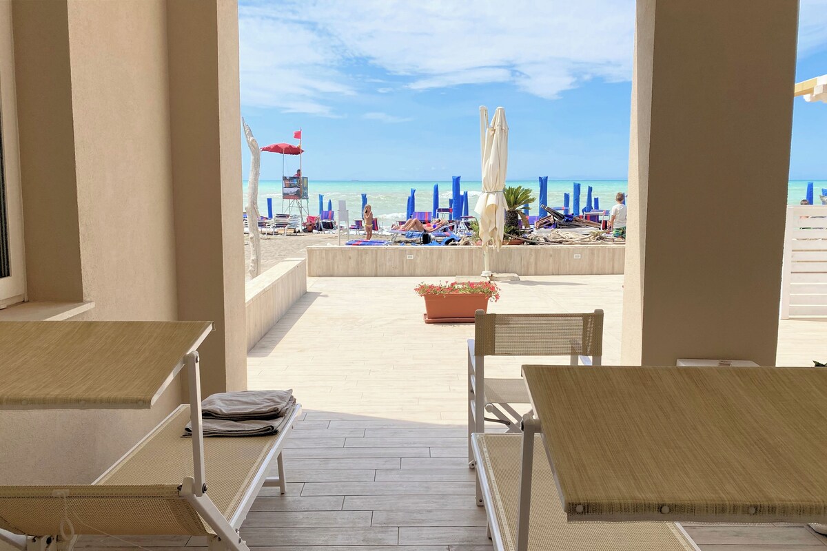 A beachside patio with two wicker chairs and a table, overlooking a beach with umbrellas and lounge chairs.
