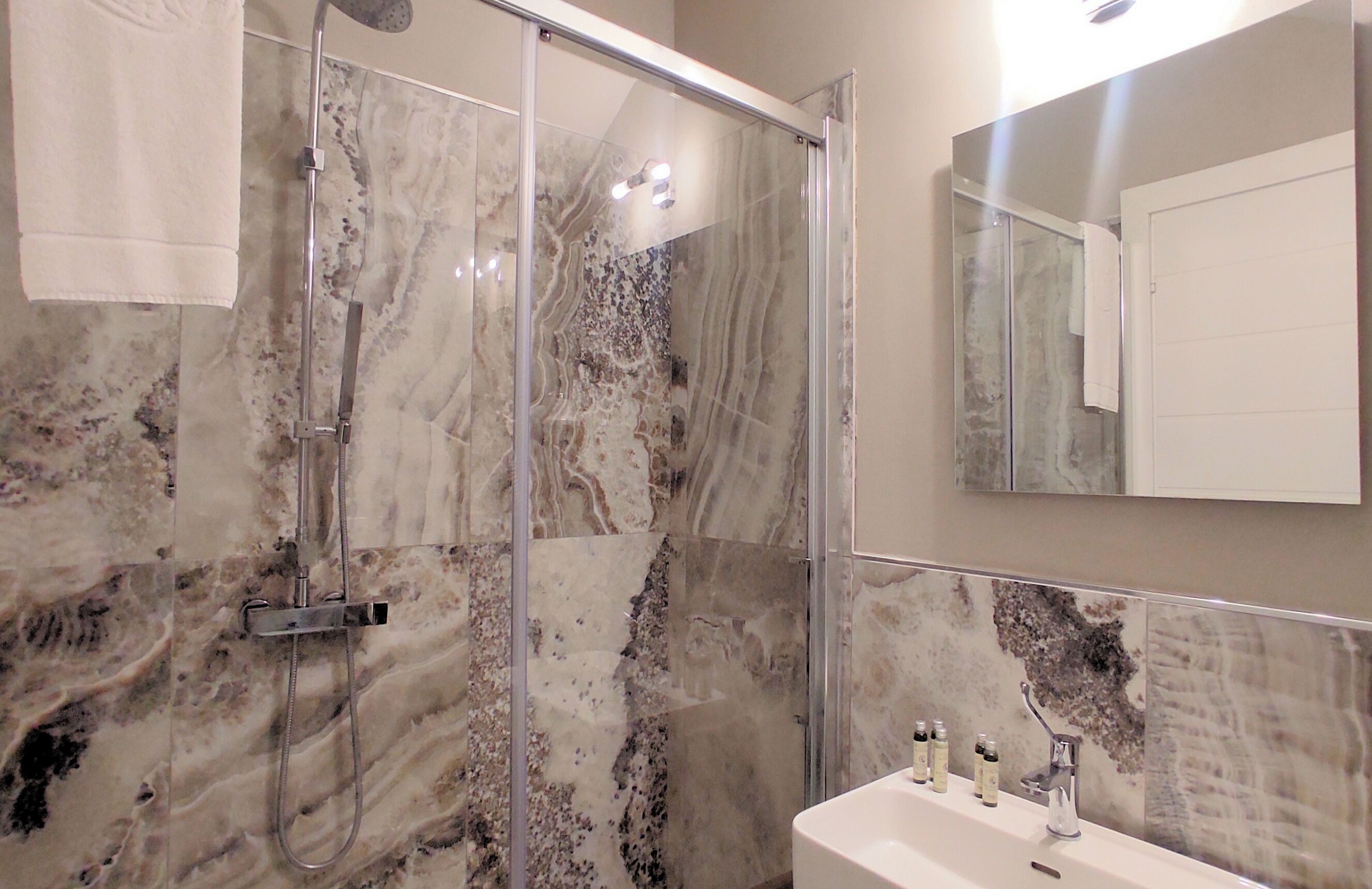 A bathroom with a glass-enclosed shower, marble walls, a sink with toiletries, and a mirror.
