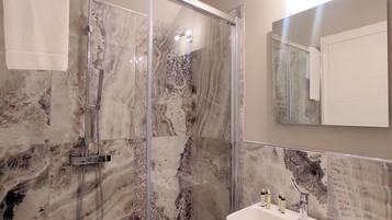A bathroom with a glass-enclosed shower, marble walls, a sink with toiletries, and a mirror.
