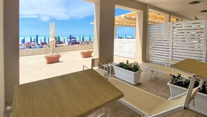 Un patio frente a la playa con mobiliario exterior, plantas en macetas y vistas al mar.