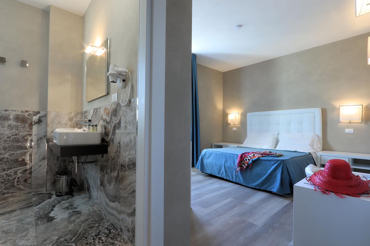 A modern bathroom with a marble sink and a separate shower area. The bedroom features a large bed with a blue bedspread and a red hat on a white nightstand.