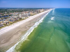 An aerial view of a coastal city with a long beach, high-rise buildings, and a clear blue sky.