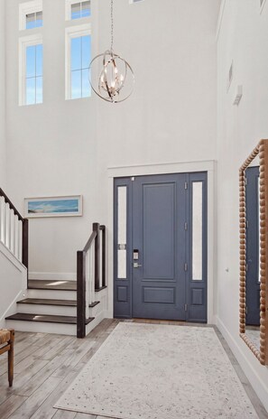 Un couloir avec un escalier, une porte bleue, un lustre et un tableau encadré au mur.