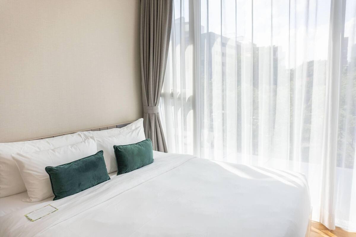 A hotel room with a large bed, white linens, and a view of the city through sheer curtains.