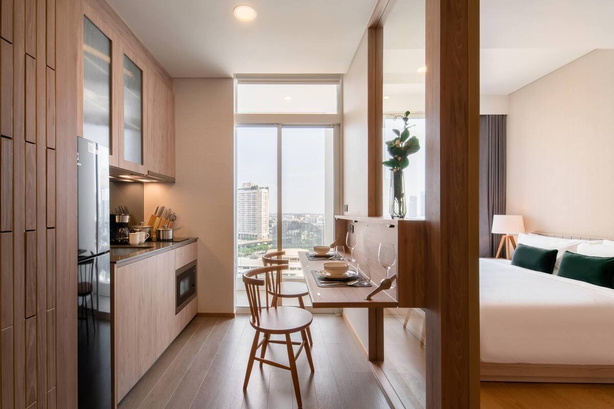 A modern hotel room with a large bed, a dining area with a table and chairs, a kitchen with wooden cabinets, and a large window offering a city view.