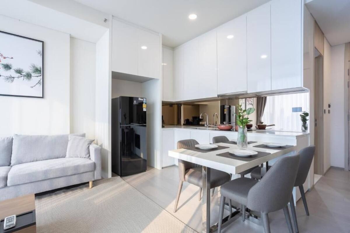A modern kitchen with white cabinets, a dining area with a white table and grey chairs, and a grey sofa in a living area.