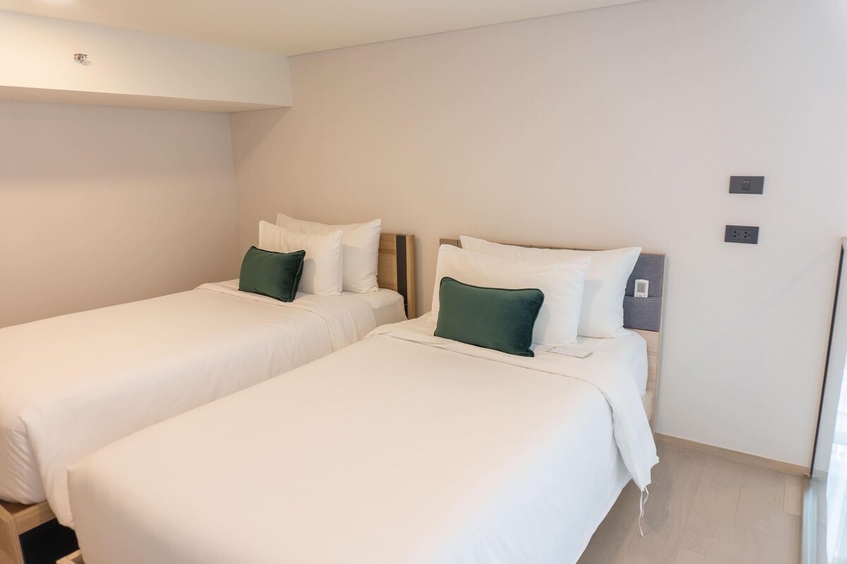 A hotel room with two single beds, each with white bedding and green pillows, a wooden headboard, and a small bedside table with a lamp.