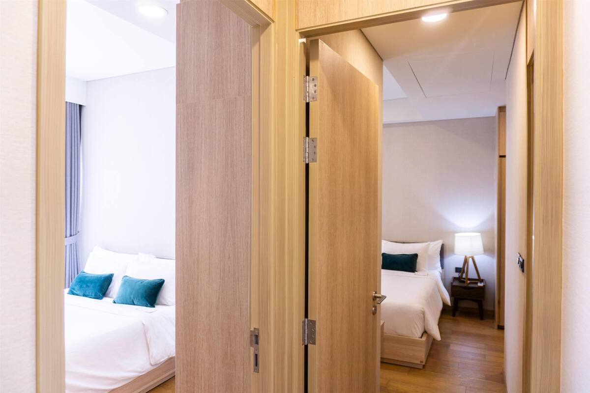 A hotel room with a wooden door, a bed with white bedding and blue pillows, a bedside table with a lamp, and a small wooden nightstand.