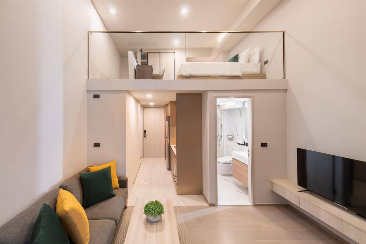 A modern apartment with a loft bedroom, a living area with a sofa and TV, and a visible bathroom.