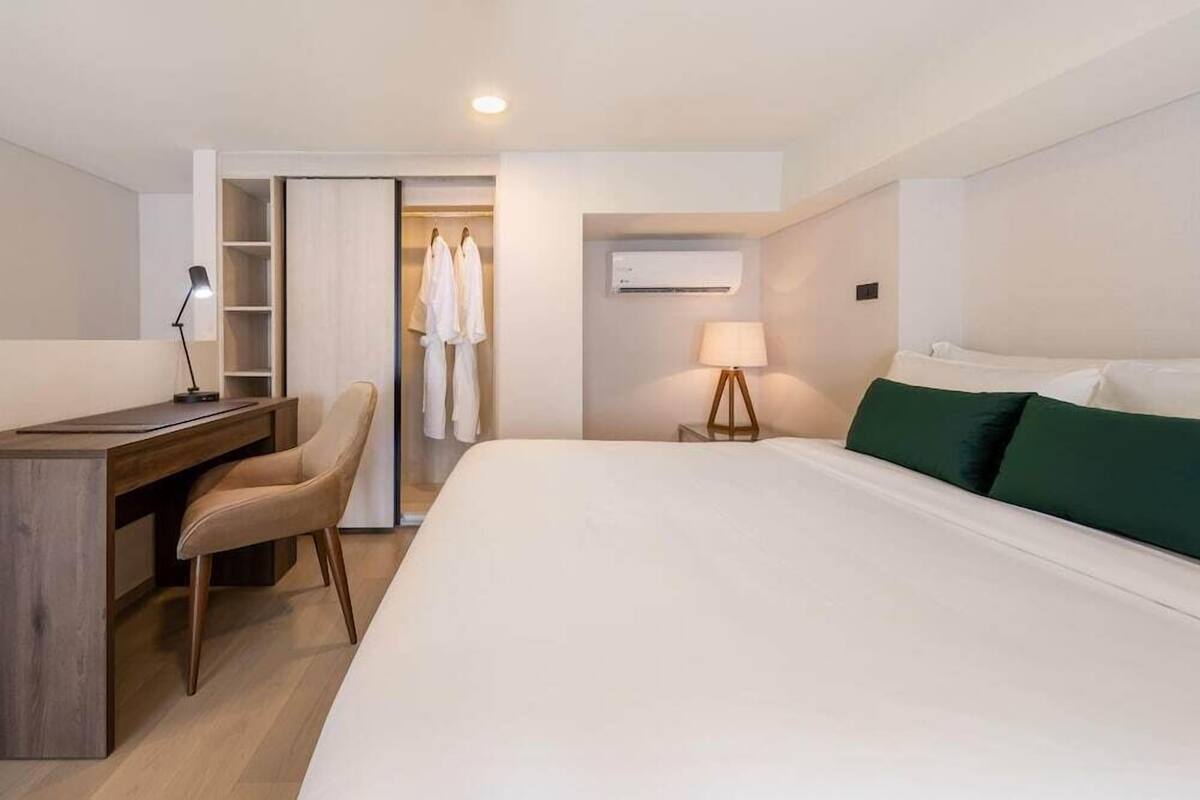 A hotel room with a bed, desk, chair, wardrobe, and a lamp.