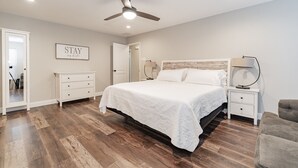 Une chambre avec un grand lit, un ventilateur de plafond, une table de chevet avec une lampe, une commode et un tableau encadré accroché au mur.