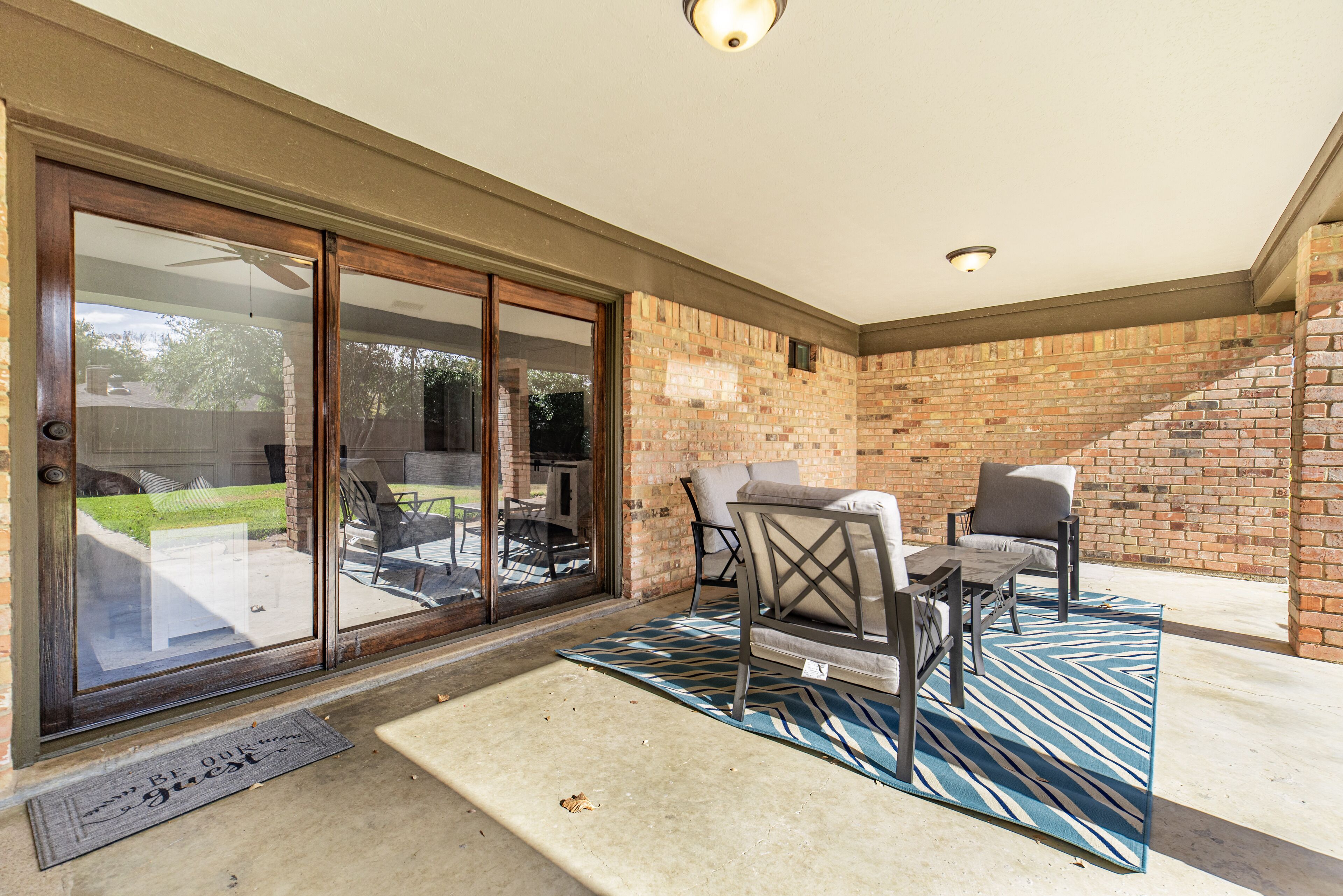 A covered patio area with a brick wall, a sliding glass door leading to a backyard, and a seating area with two chairs and a table.