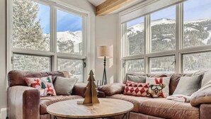 Un salón acogedor con un sofá de cuero marrón, una mesita redonda y una lámpara. Amplias ventanas ofrecen vistas a montañas cubiertas de nieve.