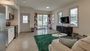 A modern kitchen with white cabinets and a microwave, a dining area with a table and chairs, a green rug, a TV on a stand, and a view of the outside through large windows. - Urban Oasis Spacious King Bed Free Parking (Nashville)