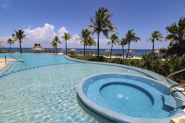 Una piscina cristalina con un jacuzzi, rodeada de palmeras y sombrillas de playa, con vista al océano.