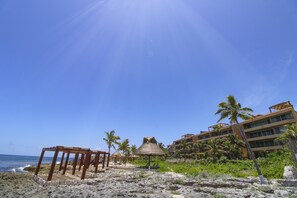 Una playa con una estructura de madera, un paraguas de paja y un edificio con varios balcones y palmeras.