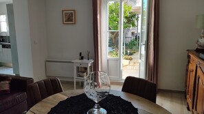 A dining area with a table set for two, a glass vase on the table, a chair, a small white side table with a phone and a book, and a view of a garden through a glass door.