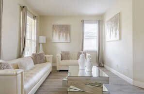 Un salon moderne avec un canapé blanc, une table basse en verre et deux fenêtres avec des rideaux.
