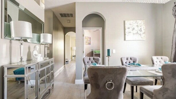 A dining area with a glass table, cushioned chairs, and a mirrored console with lamps.