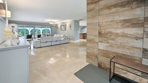 A spacious living area with a modern design, featuring a large sofa set, a wooden accent wall, and a view through large windows. - Luxurious Naples Home. Heated Pool. Mins to Beach. (Naples)