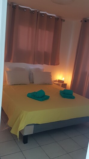 A neatly made bed with two pillows and two folded towels, a bedside table with a lamp, and a window with curtains. - Anse à l'âne - Spacious T2 - Exceptional view (Les trois ilets)