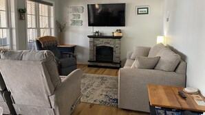 A living room with a stone fireplace, two grey sofas, a wooden coffee table, and a flat-screen TV mounted on the wall. - Sunrise Lake House, Comfort-Hot Tub-Beach! (Saint Albans)