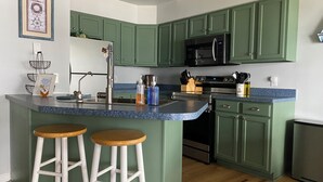 A kitchen with green cabinets, a stainless steel refrigerator, a microwave, and a sink. There are two wooden stools at the island. - Sunrise Lake House, Comfort-Hot Tub-Beach! (Saint Albans)
