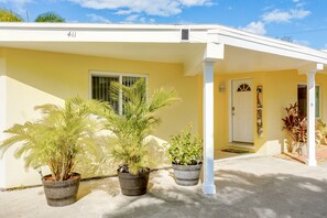 Une maison jaune avec un porche blanc, le numéro 411, et trois plantes en pot devant.