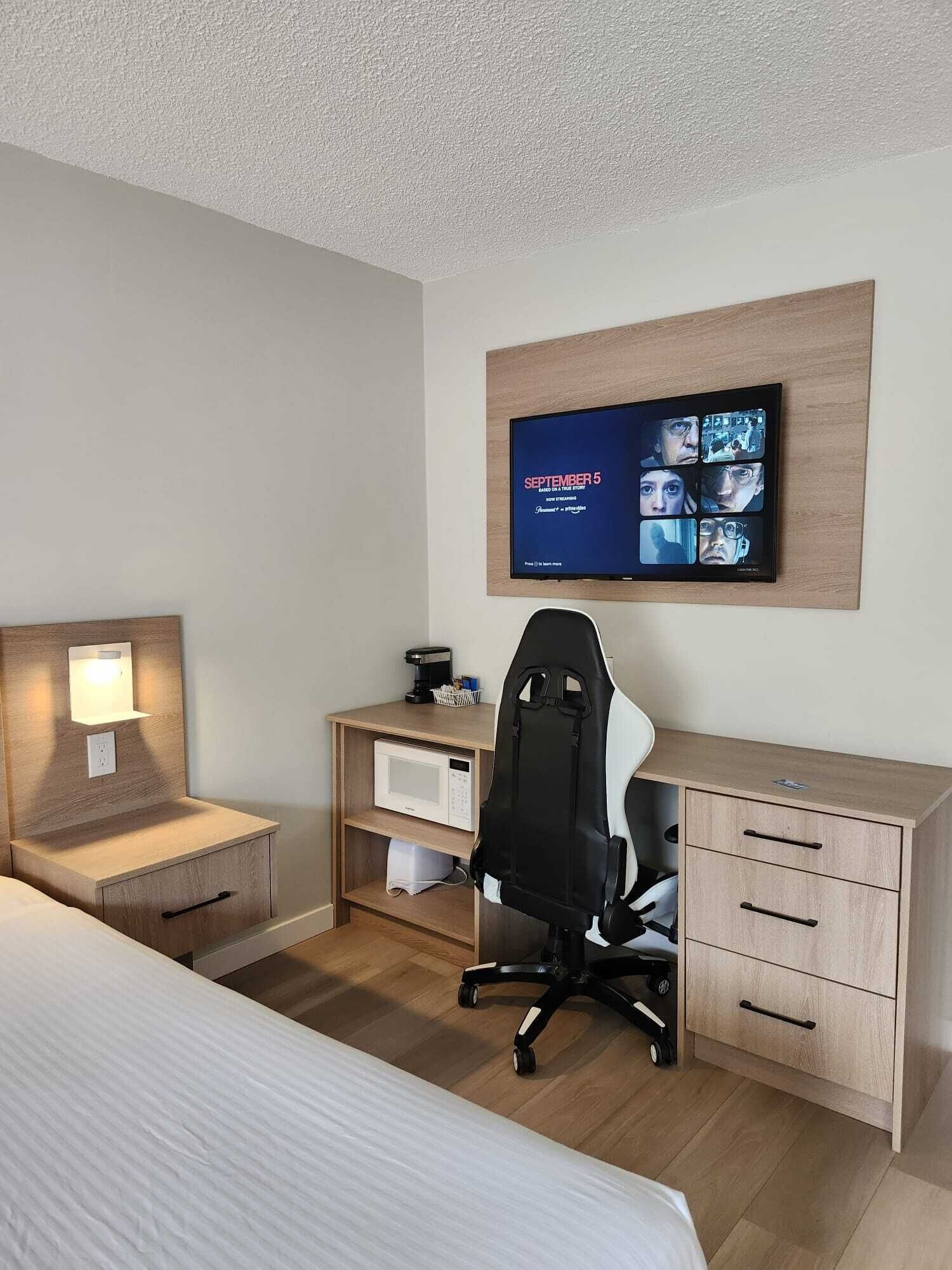 A hotel room with a bed, a desk with a computer, a chair, a TV mounted on the wall, and a small shelf with a lamp.