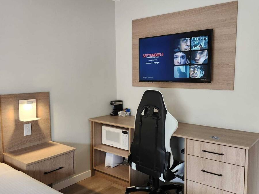 A hotel room with a bed, a desk with a computer, a chair, a TV mounted on the wall, and a small shelf with a lamp.